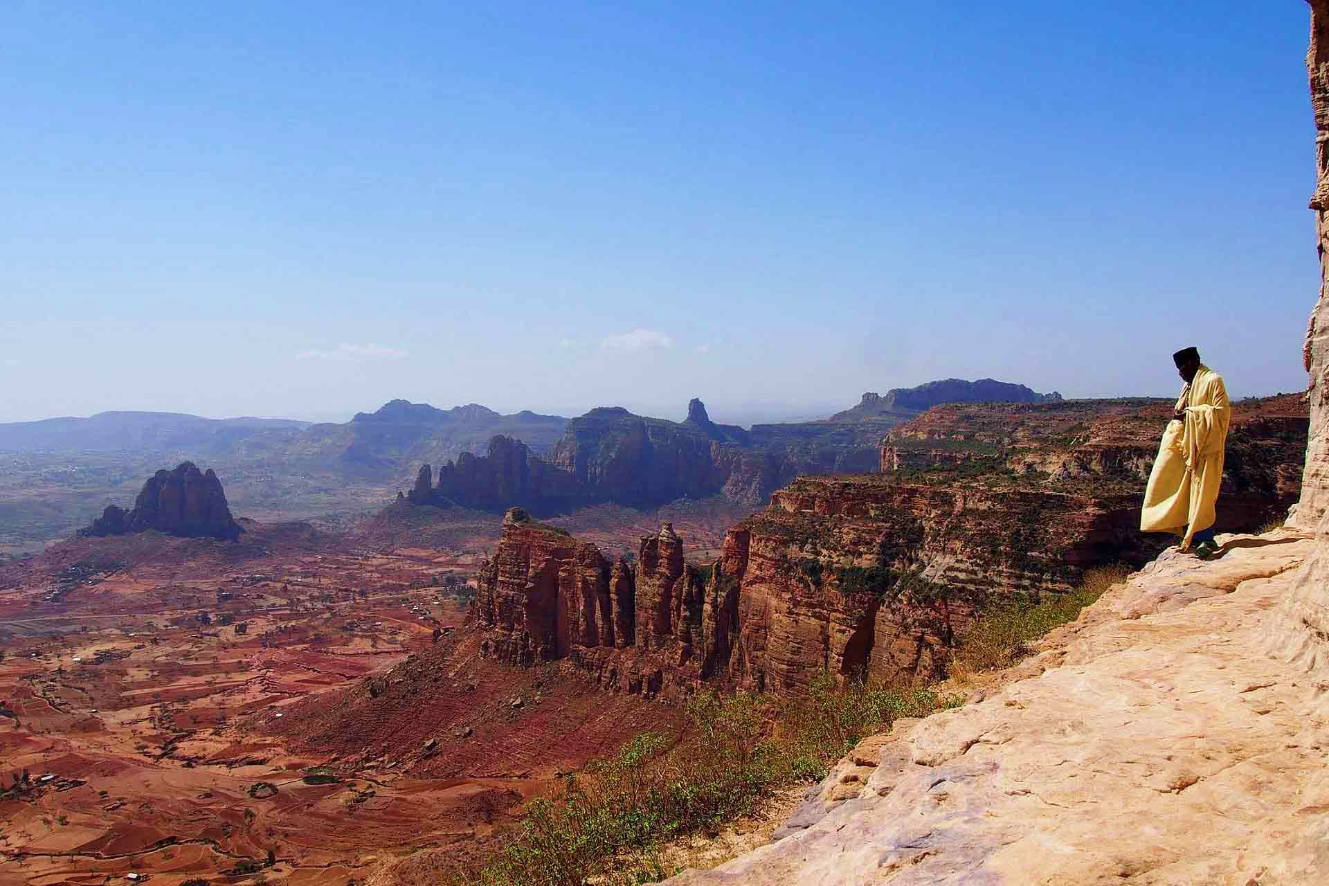 Monk in yellow robe standing on cliff overlooking dramatic rock formations and valley landscape in Ethiopia's Simien Mountain